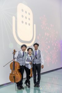Noah Lee, with his cello, poses next to his fellow musicians in front of a From the Top logo projected on a white wall.